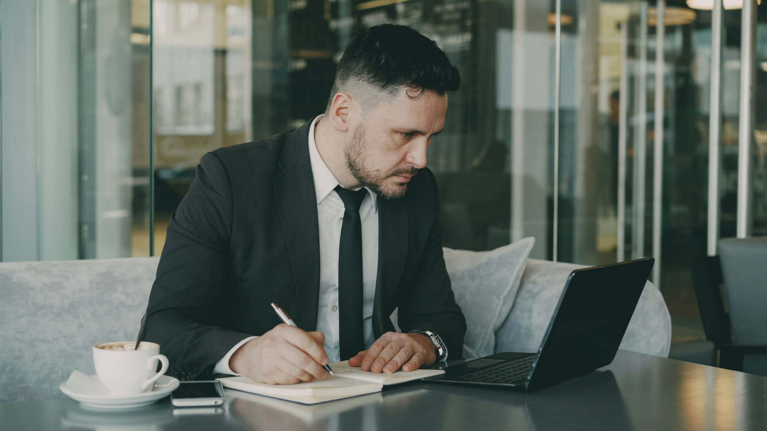 A man in a suit sits at a table, writing in a notebook while working on a laptop in an office setting.