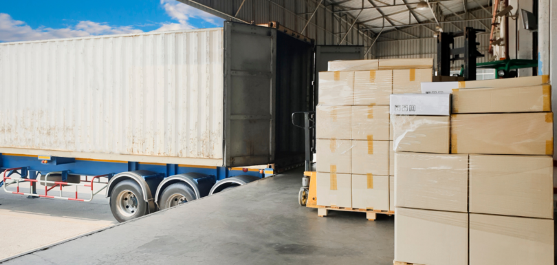 Pallets of packaged goods beside an open freight container at a warehouse